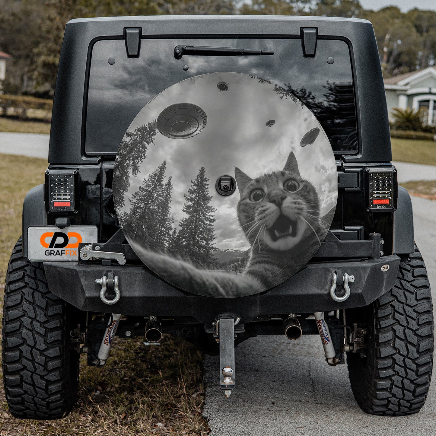a black jeep with a large, round spare tire cover on the rear window. The cover depicts a cat with a surprised expression, looking at the camera.