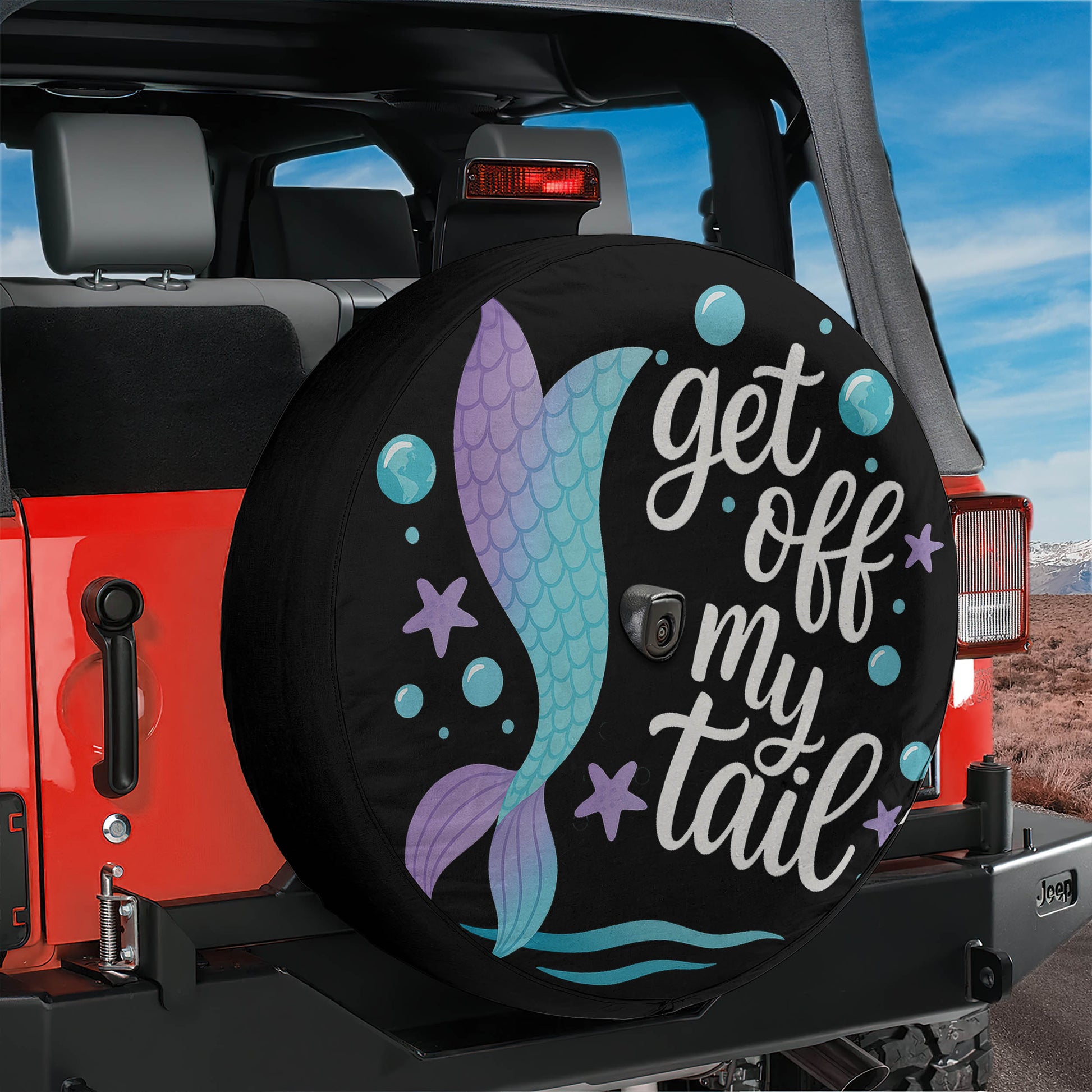 A black tire cover with a mermaid design and the text "get off my tail" printed on it, attached to the rear of a red Jeep.