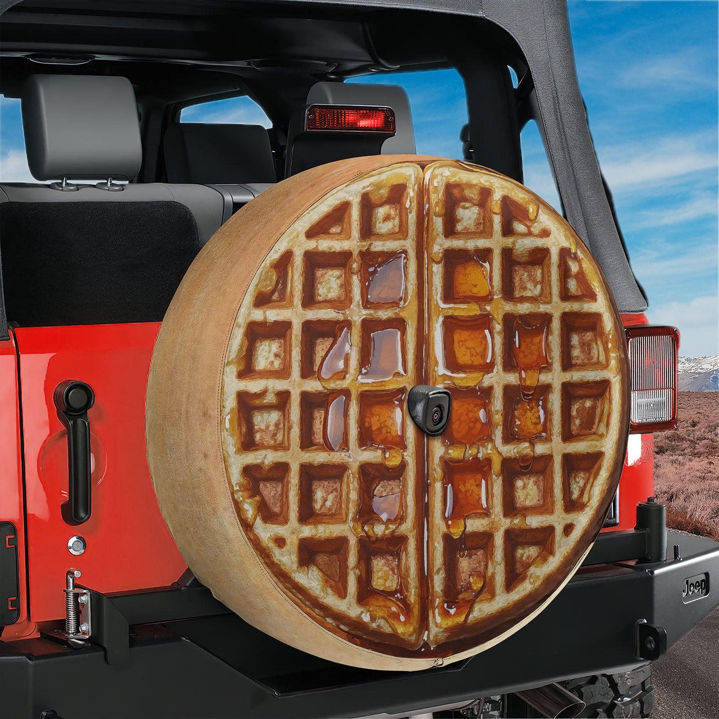A waffle-shaped spare tire is attached to the rear of a red Jeep, with a scenic landscape in the background.