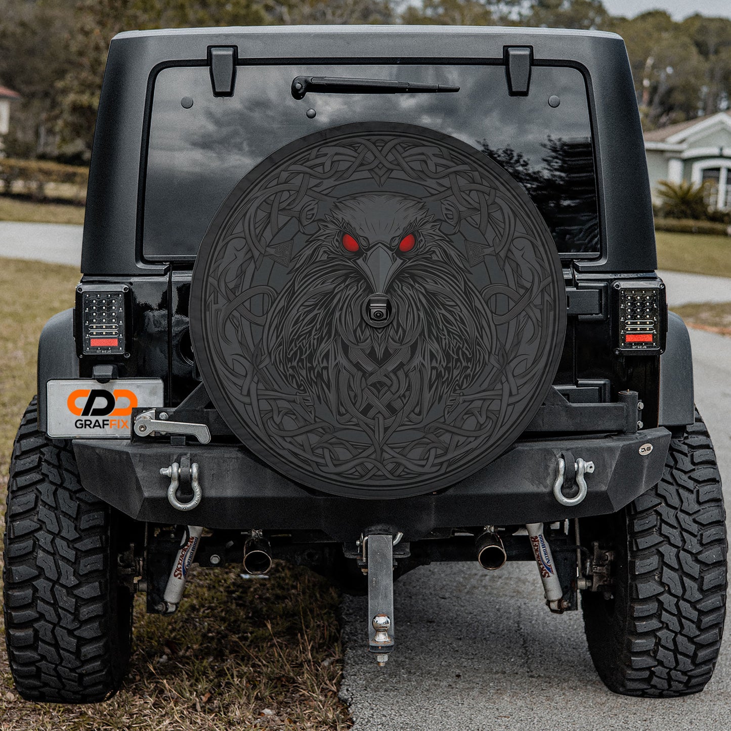 a black Jeep Wrangler with a large, decorative wheel cover featuring a fierce-looking wolf or wolf-like creature design. The Jeep is parked on a road, and there is a house visible in the background.