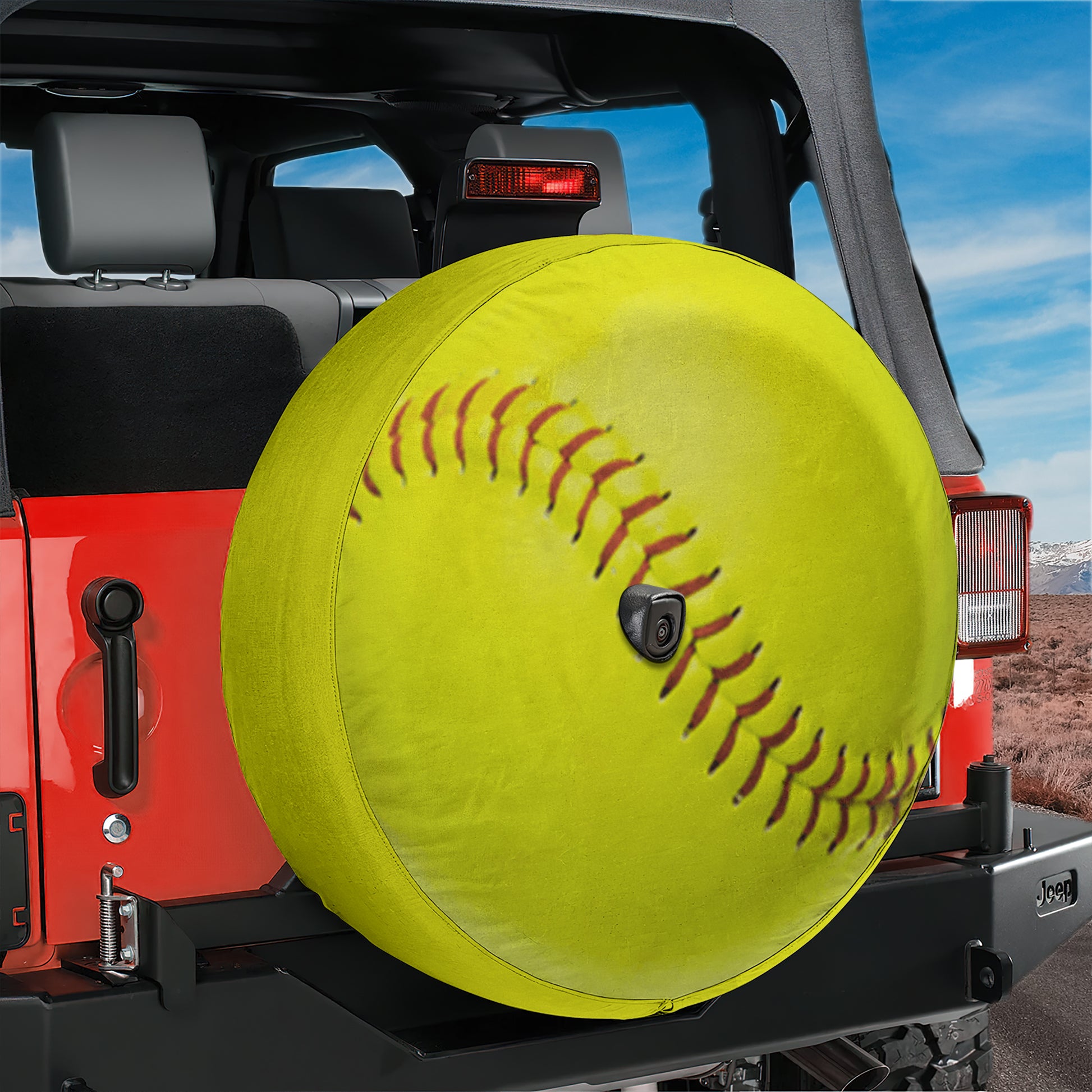 A large yellow softball with red stitching is mounted on the rear of a red Jeep, with a clear blue sky in the background.