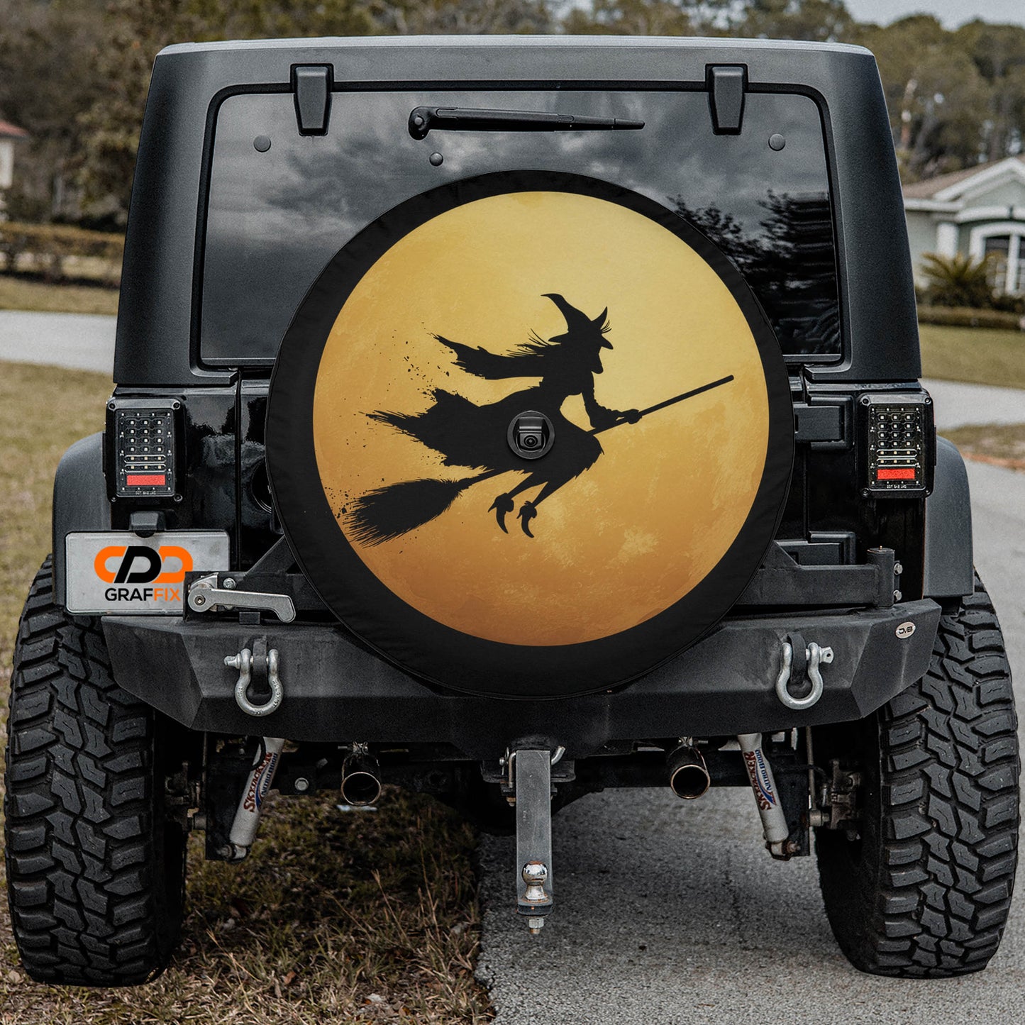 a black Jeep Wrangler with a large tire cover featuring a witch on a broomstick. The cover has a yellow background and black silhouette of the witch.