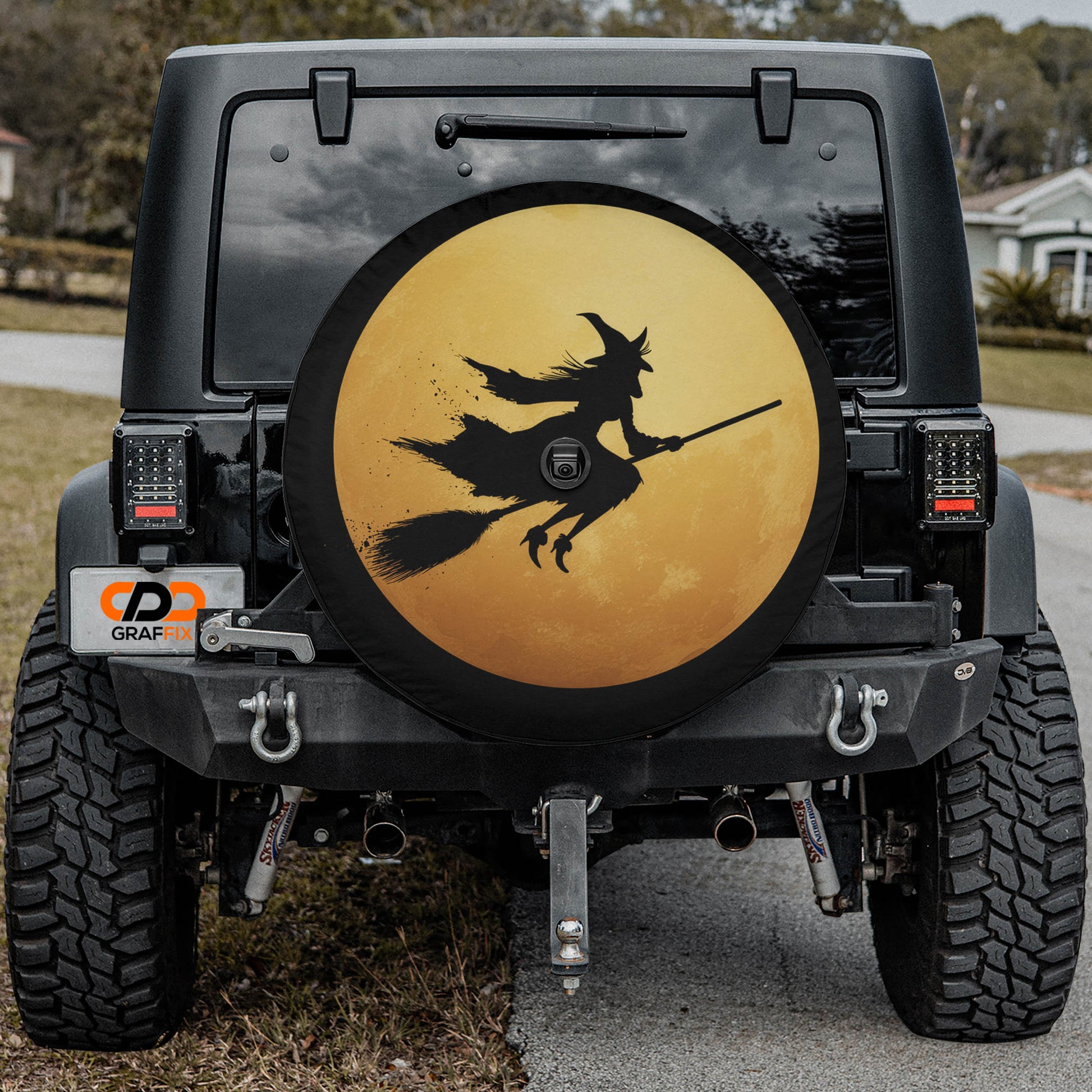 a black Jeep Wrangler with a large tire cover featuring a witch on a broomstick. The cover has a yellow background and black silhouette of the witch.