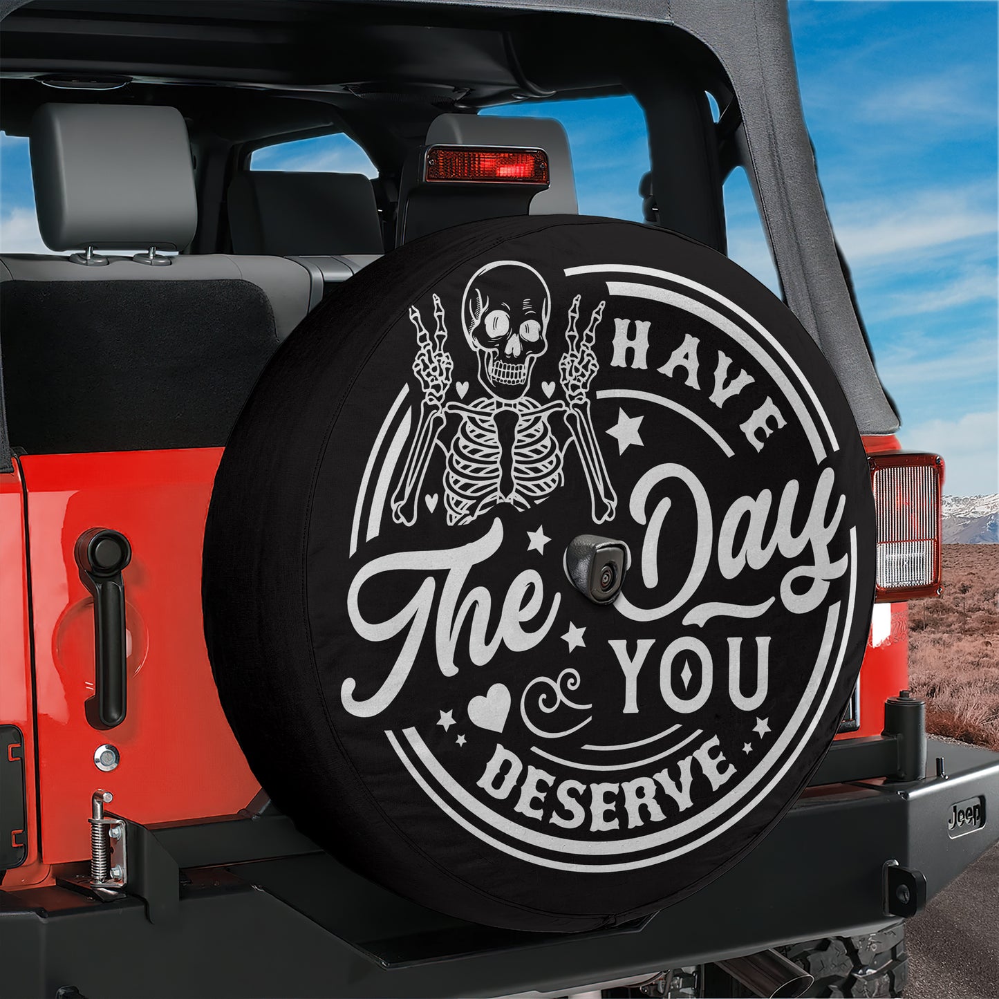 A black tire cover with a skeleton and the text "Have a Day, You Deserve" printed on it, mounted on the rear of a red Jeep.