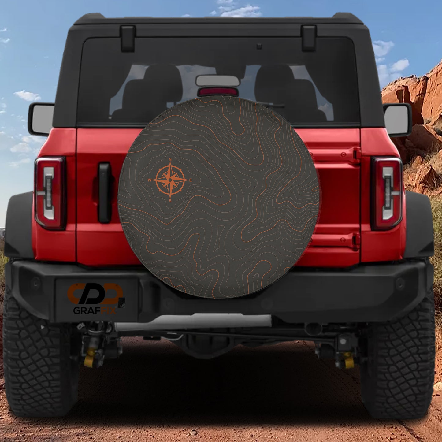 A red Jeep Wrangler with a large off-road tire cover featuring a compass design, parked on a dirt road with a rocky hill in the background.