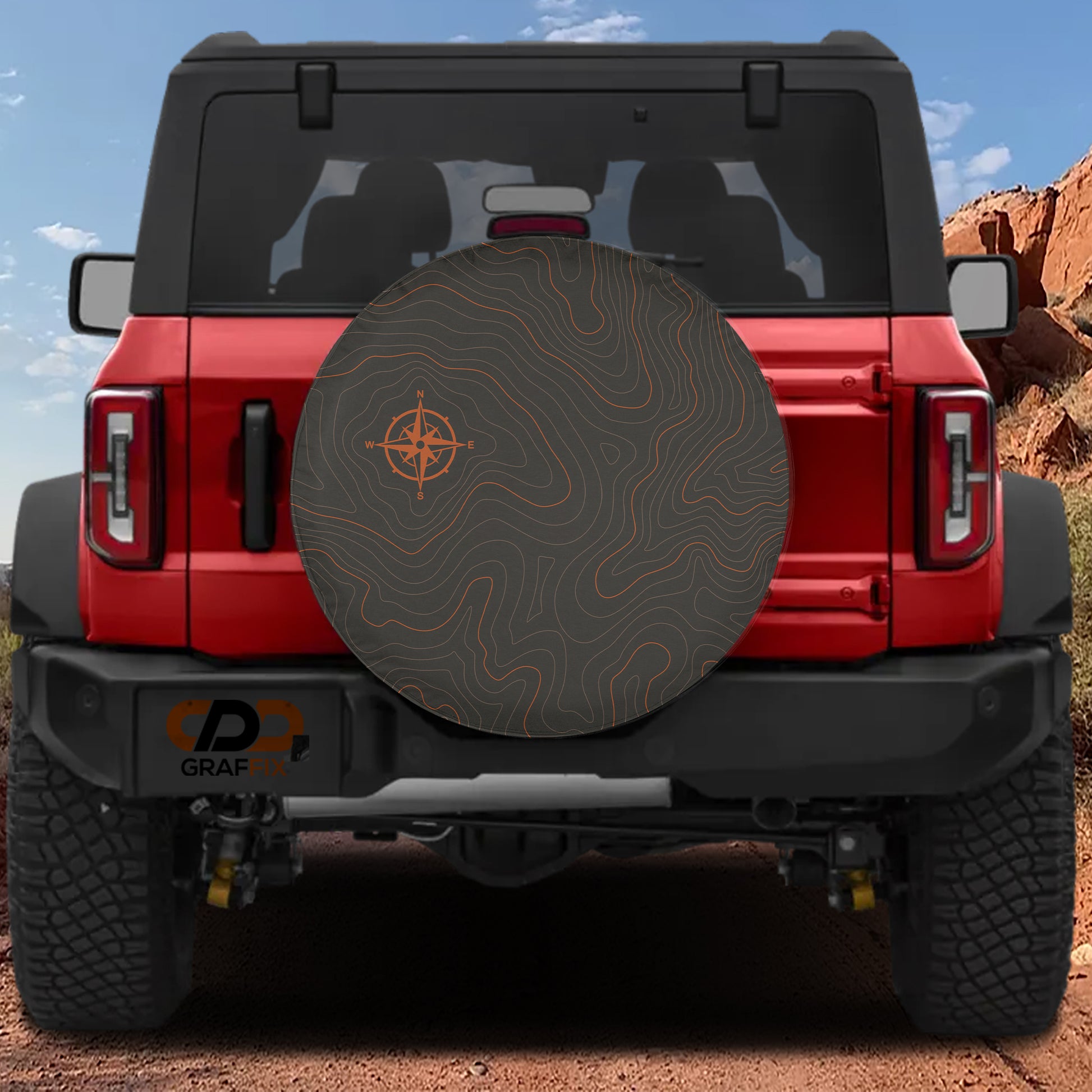 A red Jeep Wrangler with a large off-road tire cover featuring a compass design, parked on a dirt road with a rocky hill in the background.