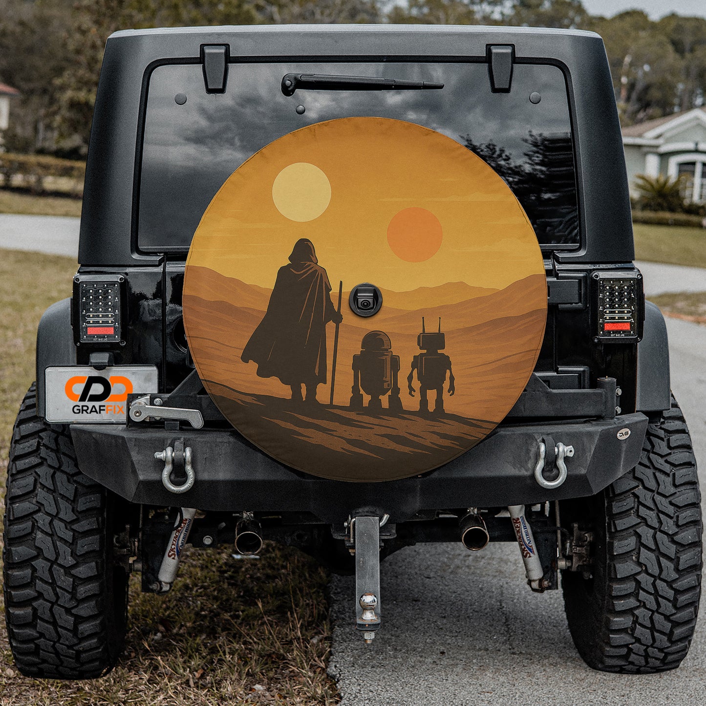 a Jeep Wrangler with a large, round spare tire cover on the rear of the vehicle. The cover displays a desert scene with a silhouetted figure and two robot-like figures.