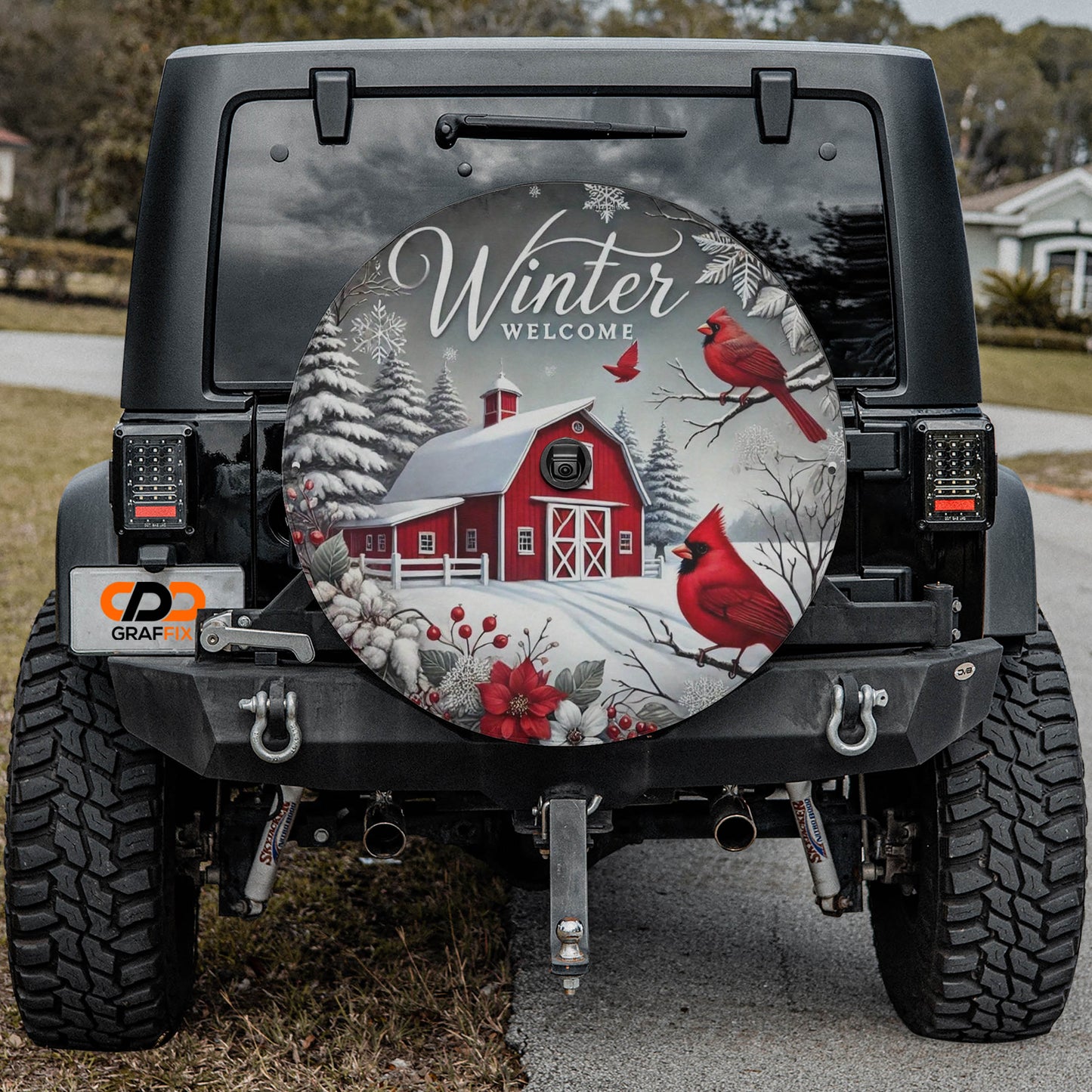 a black jeep with a large winter-themed spare tire on the rear. The spare tire has a design depicting a red barn, snow-covered trees, and two red birds perched on branches.