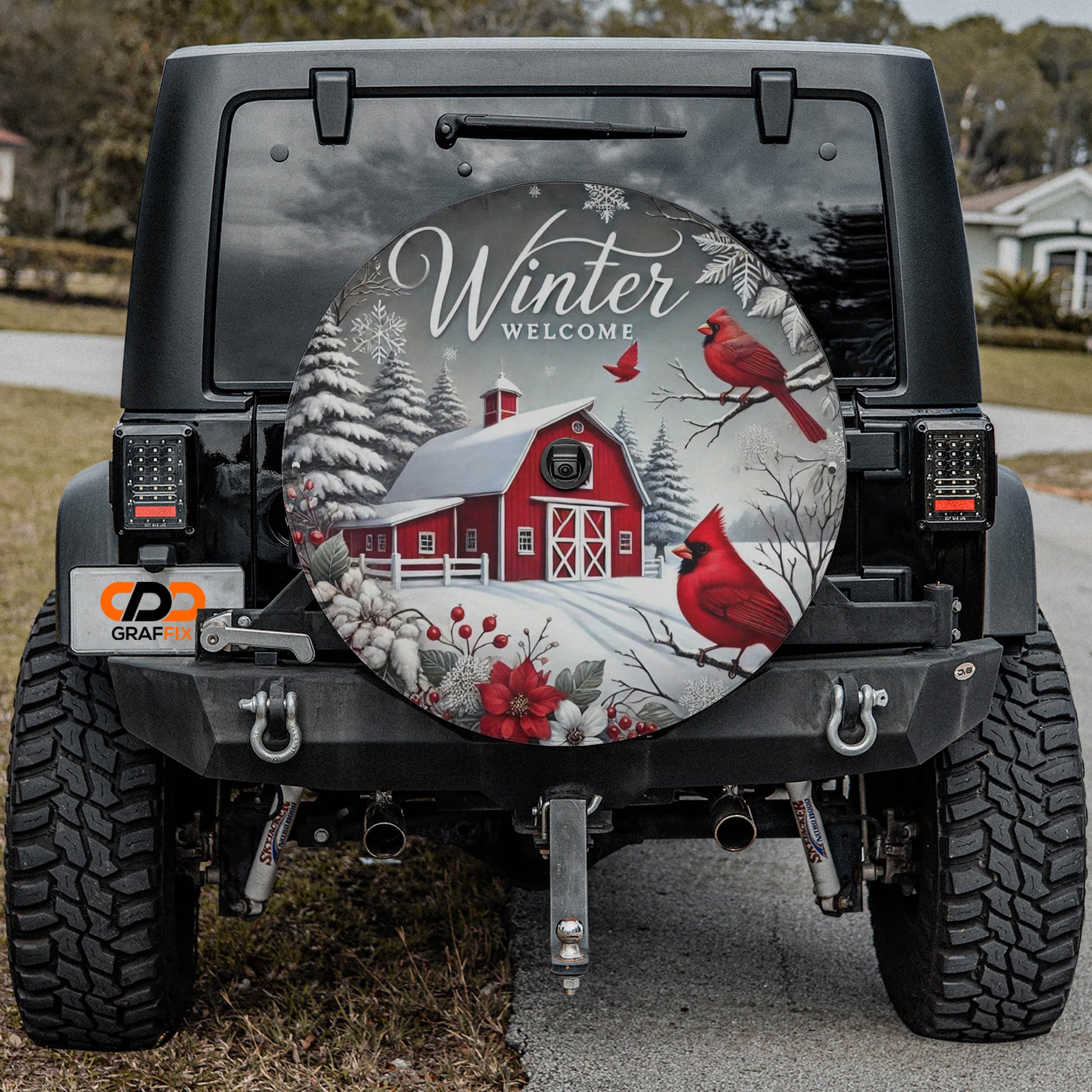 a black jeep with a large winter-themed spare tire on the rear. The spare tire has a design depicting a red barn, snow-covered trees, and two red birds perched on branches.