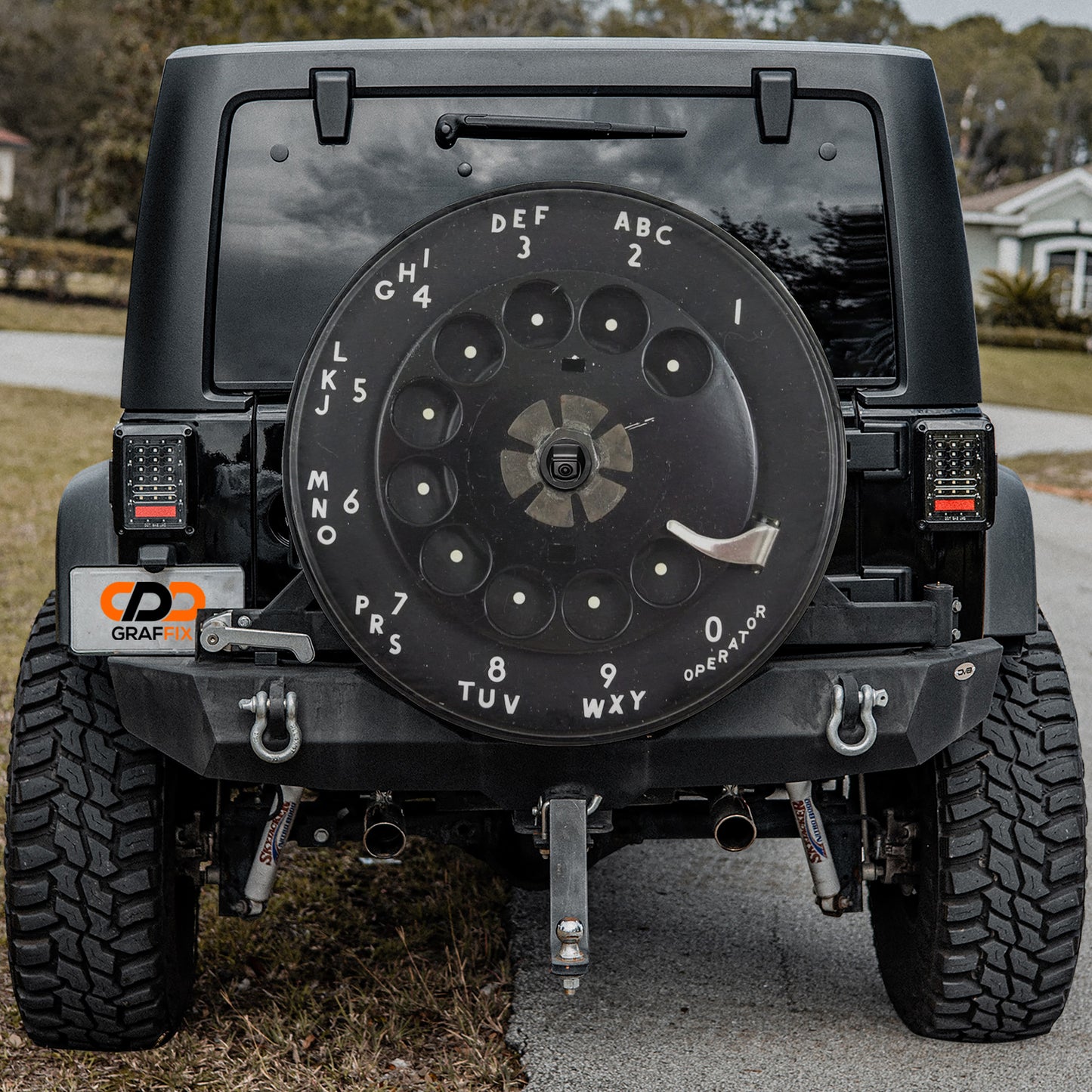 a black Jeep Wrangler with a large black wheel cover on the rear of the vehicle. The wheel cover has numbers and letters printed on it, indicating a dial-like design.