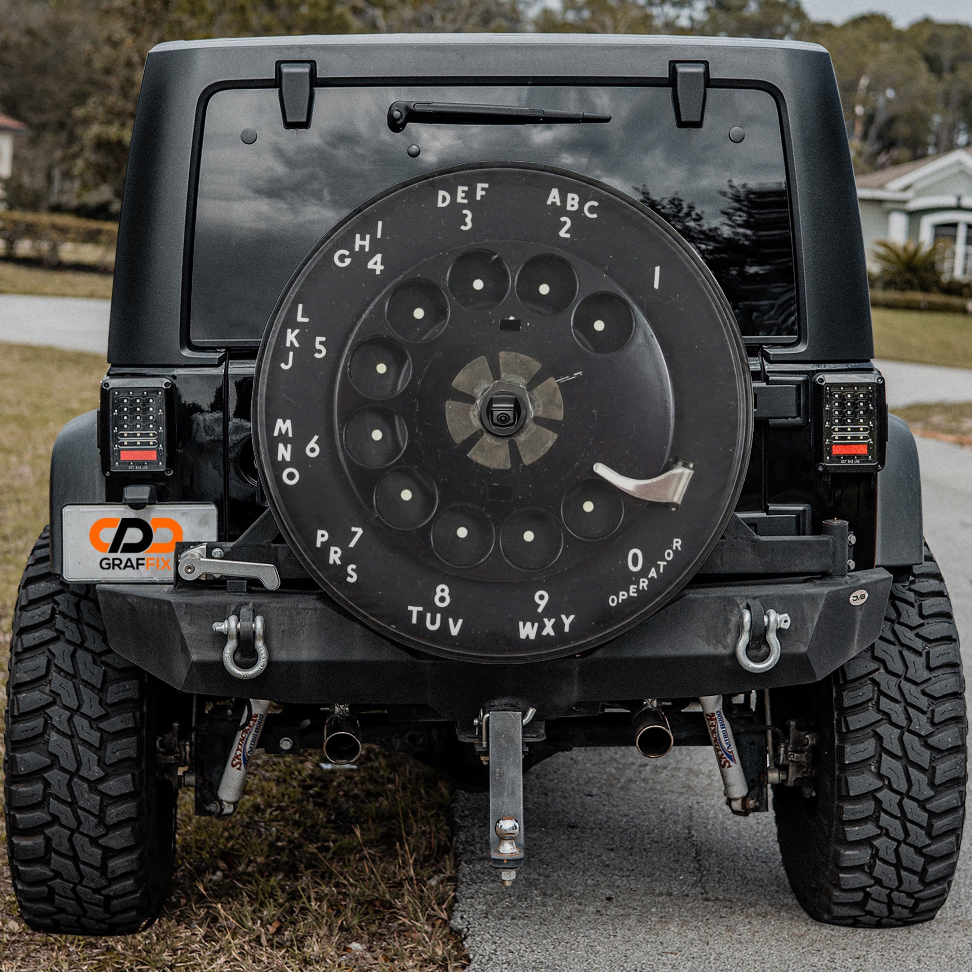 a black Jeep Wrangler with a large black wheel cover on the rear of the vehicle. The wheel cover has numbers and letters printed on it, indicating a dial-like design.