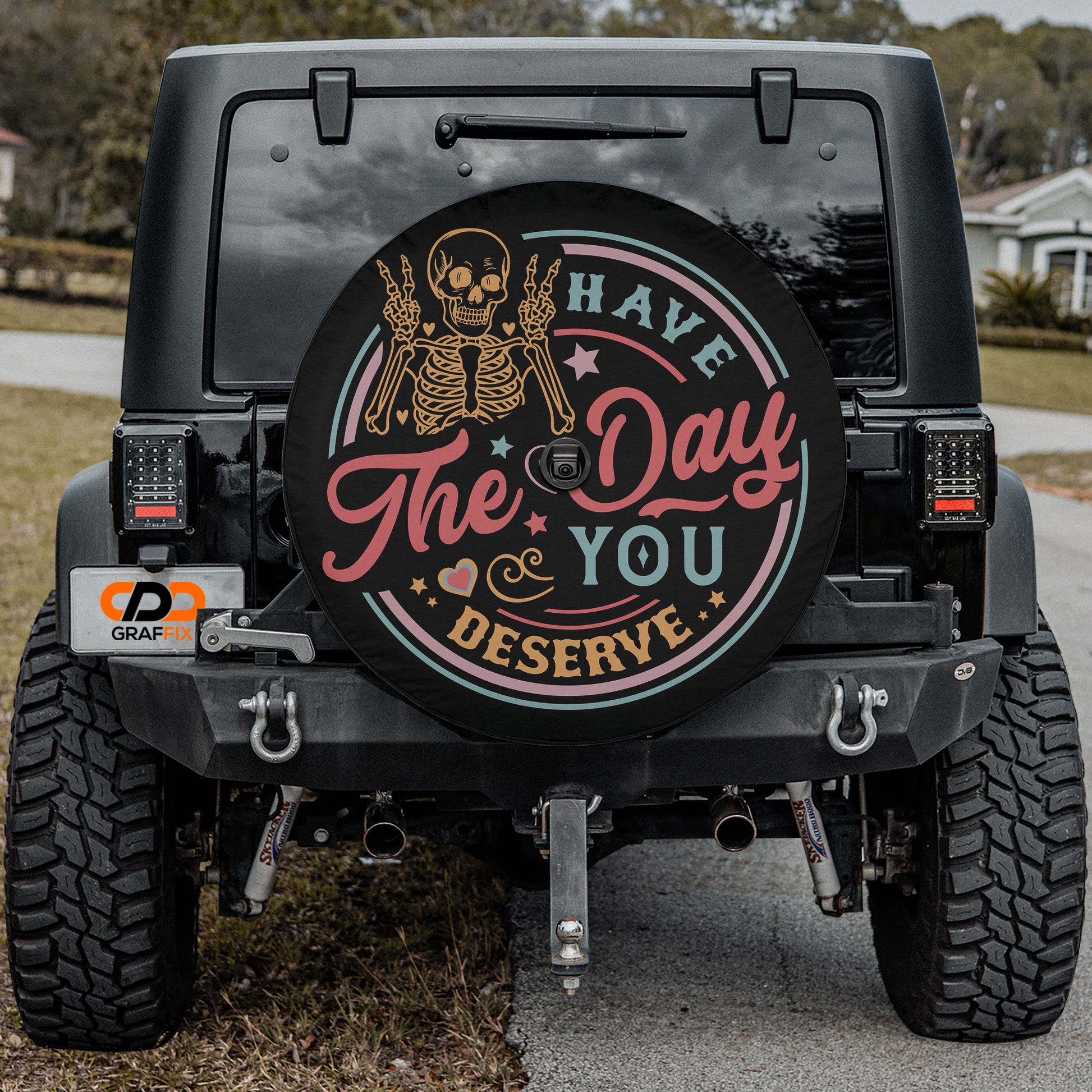 the back of a black Jeep Wrangler with a custom license plate and a wheel cover featuring a skeleton and the text "The day you deserve".