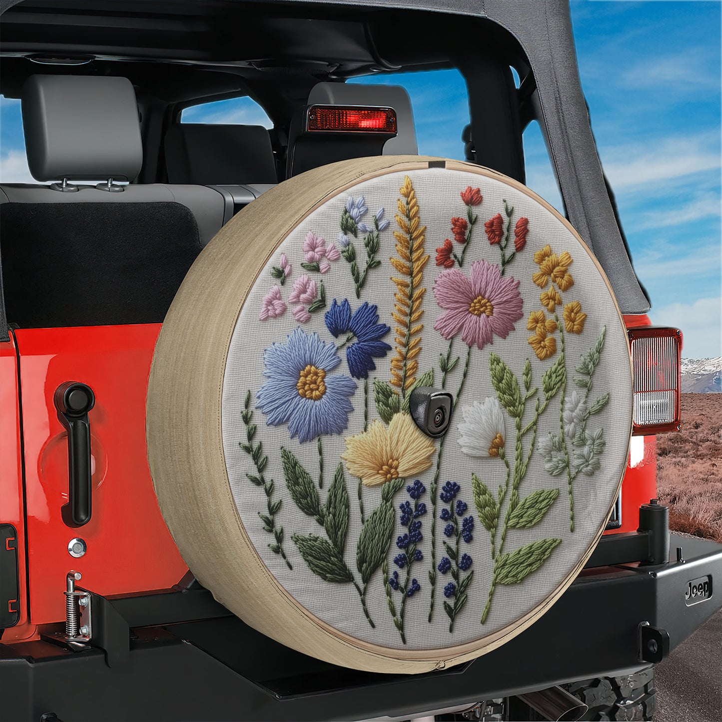 A colorful wheel cover with embroidered flowers and leaves is attached to the rear of a red Jeep vehicle.