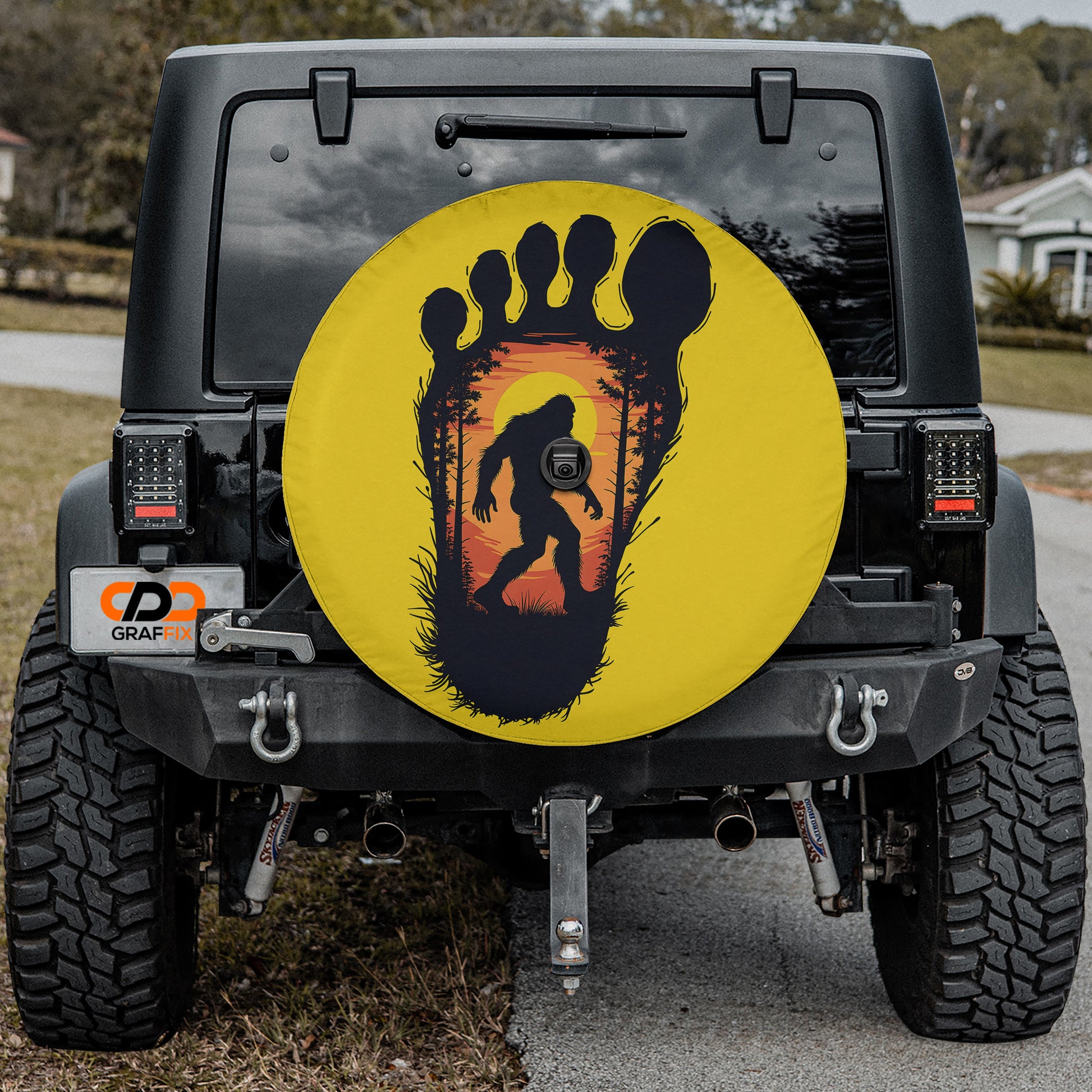 a black Jeep Wrangler with a large tire cover on the rear of the vehicle. The cover has a silhouette of a Sasquatch, a mythical creature, and a sunset in the background.